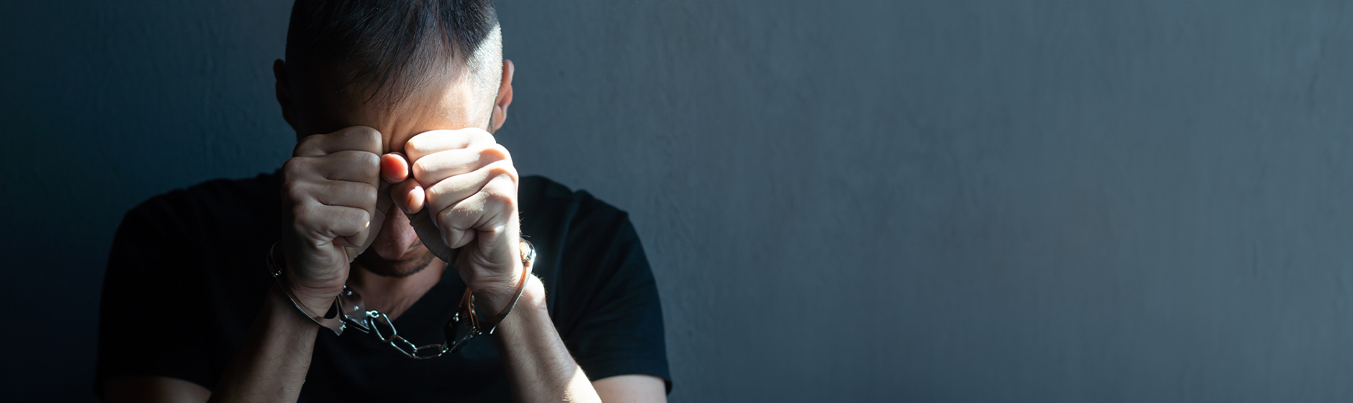 A person wearing a black shirt is in handcuffs, sitting against a dark background, embodying the weight of their circumstances. With their head bowed and hands clenched, shadows create a somber silhouette as they await hope from a criminal defense lawyer.