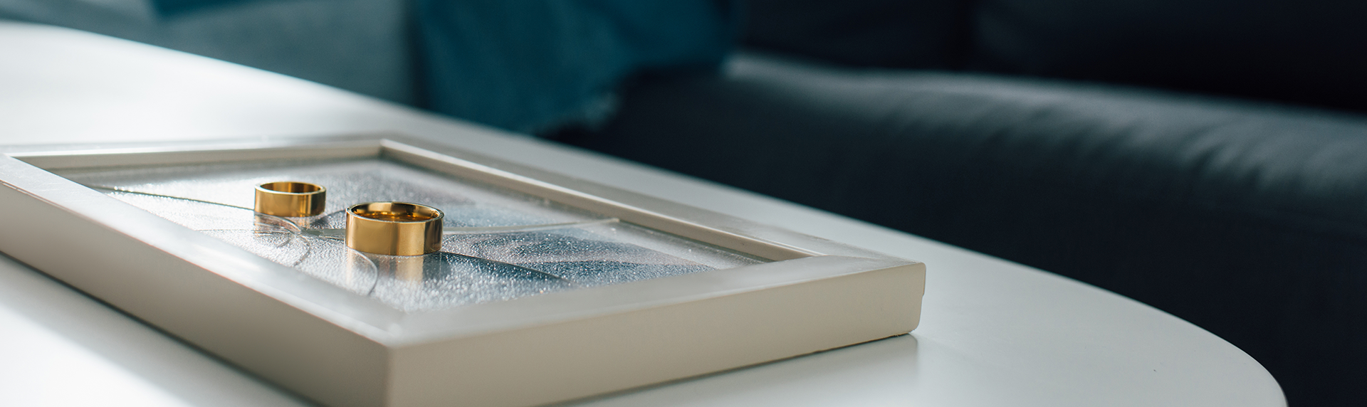 Two gold rings rest on a rectangular, glass-covered surface with visible cracks, placed on a sleek white table. A dark blue sofa is blurred in the background, creating a modern and minimalist setting—much like the poised precision of a criminal defense lawyer's office. Morris County Divorce Lawyers concept image