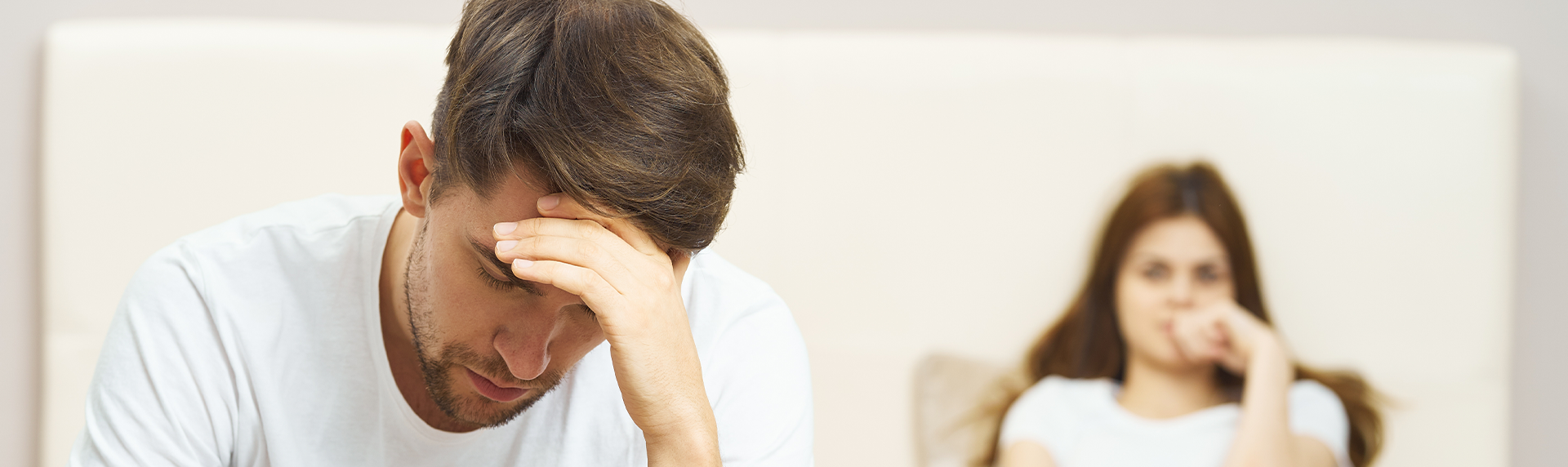 A man sits on a bed with his head in his hand, looking distressed, as if contemplating calling a criminal defense lawyer. In the background, a woman watches him thoughtfully. Both are in casual white clothing, and the setting appears to be a bedroom.