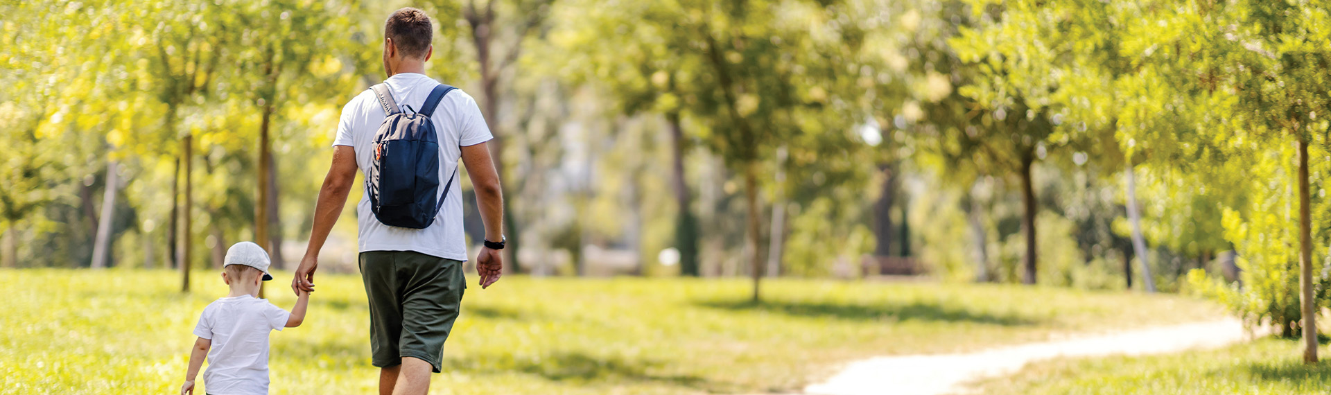 A man with a backpack walks hand-in-hand with a child wearing a hat. They stroll along a sunlit path in a park surrounded by green grass and trees, enjoying the peaceful and bright scene—a simple reminder of the joys that can come when fathers' rights are cherished and protected.