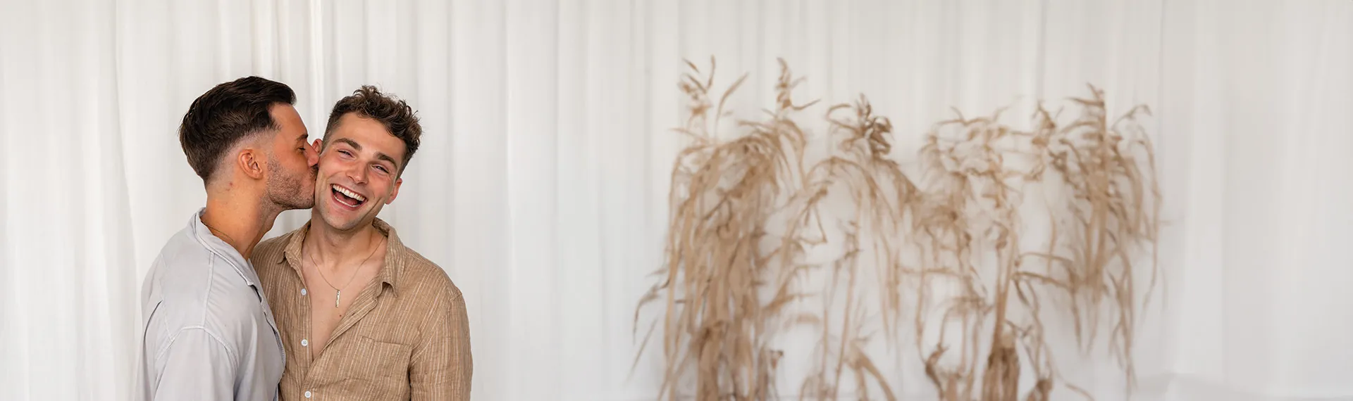 Two people, representing same-sex partners, are smiling in front of a white curtain backdrop. One person on the left is playfully kissing the cheek of the other, while decorative dry plants add a touch of charm to the background on the right.