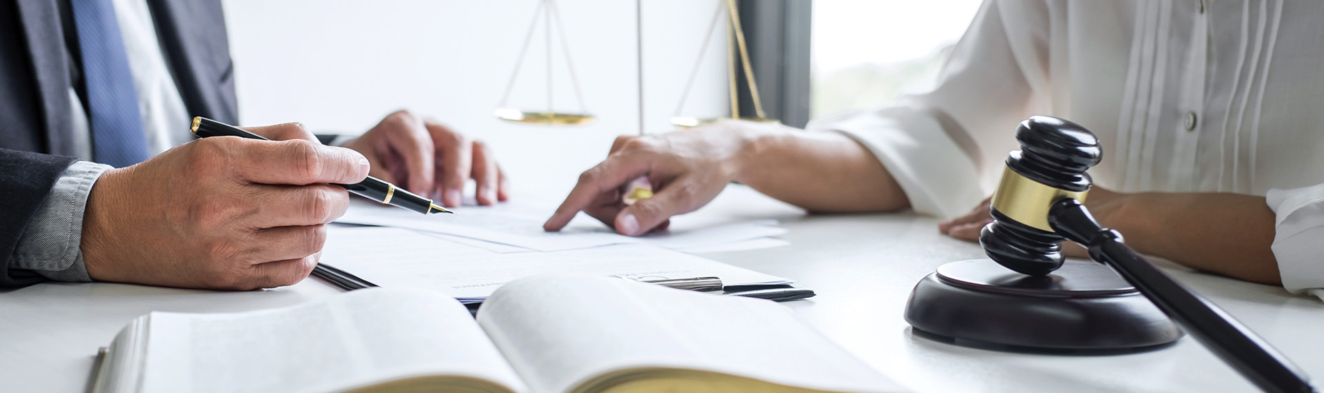 Two individuals, likely representing lawyers, are seated at a table discussing documents. One holds a pen, pointing at the papers. A gavel and scales of justice sit nearby, suggesting a legal setting. The focus is on their hands and the objects on the table.