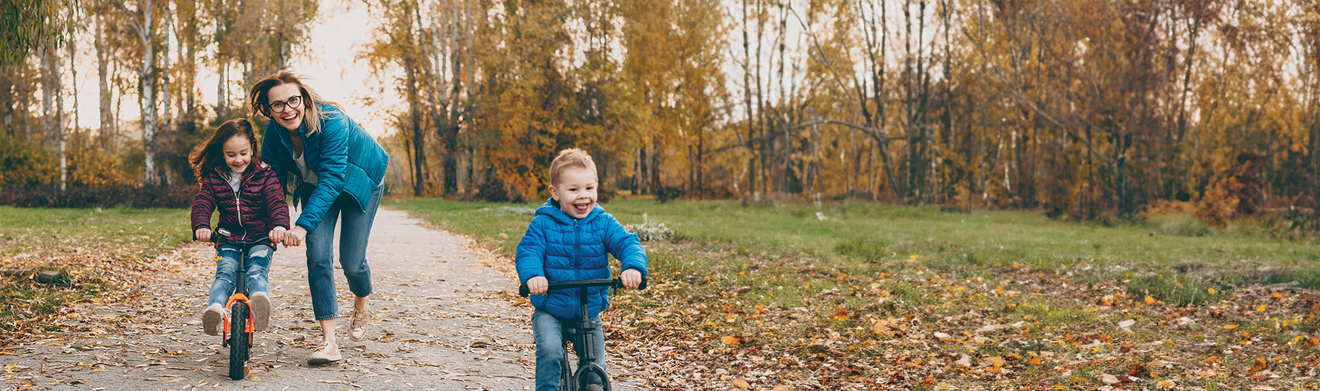 A woman joyfully running alongside two children, each riding bikes on a leafy path in a park. The vibrant autumn colors of the trees create a warm and lively atmosphere, echoing the positive vibe of their cherished moments together.