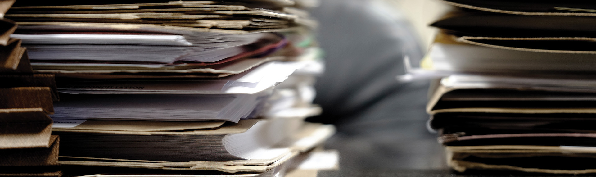 Stacks of assorted documents and files, including prenuptial agreement help papers, are piled high on a desk, with papers peeking out from folders. The scene suggests a busy and cluttered workspace, with a blurred figure in the background adding to the sense of activity.