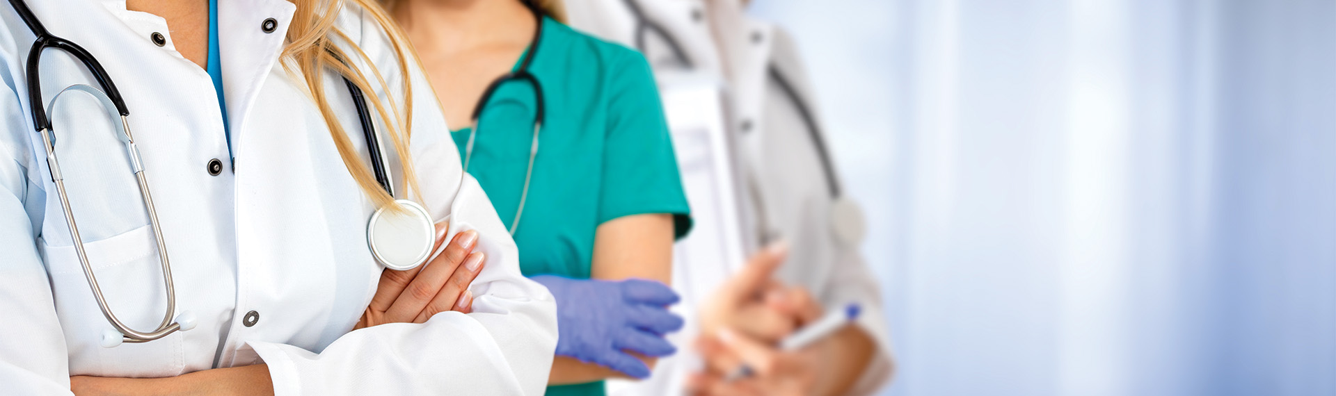 Three doctors in white coats and scrubs stand side by side. One is wearing a stethoscope, another has gloves, and the third is holding a pen. They are in a brightly lit environment.