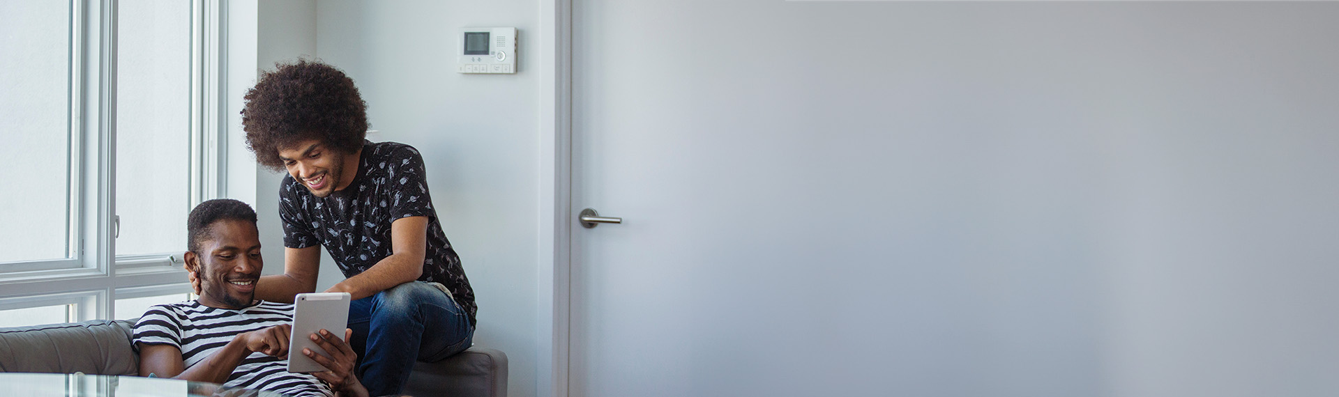 Two people are sitting on a couch in a bright room, smiling and looking at a tablet. One wears a striped shirt, the other a patterned one, perhaps browsing for same-sex prenuptial agreement attorneys. A thermostat is on the wall behind them, with a door to the right.