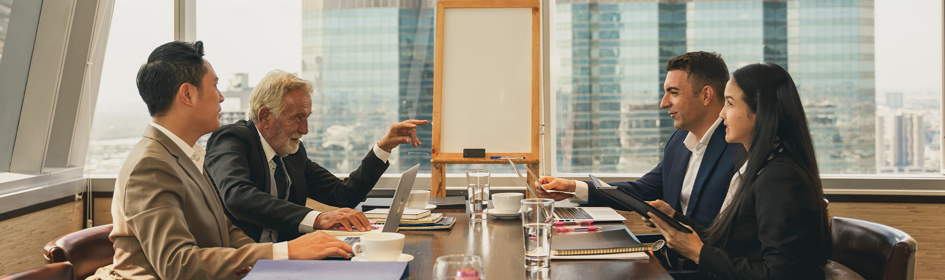 A group of four people in business attire are having a meeting focused on the benefits of mediation, seated around a table with laptops, papers, and glasses of water. Large windows light the room as an empty easel stands in the background.