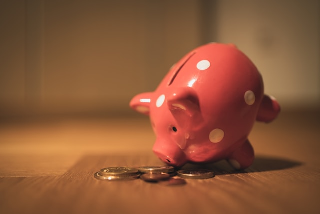A red piggy bank with white polka dots is tipped over on a wooden surface, with scattered coins—a playful reminder to share your retirement savings—visible in front of it.