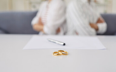 Two gold rings on a table, with a pen and paper in the background. Two people, blurred and sitting back-to-back, suggest tension. Focus is on the rings, conveying themes of separation or divorce.