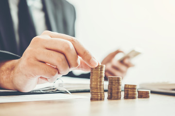 A person in a suit stacks coins into ascending piles on a desk, symbolizing financial growth or investment, while managing their post-divorce budget on a mobile phone in the other hand.