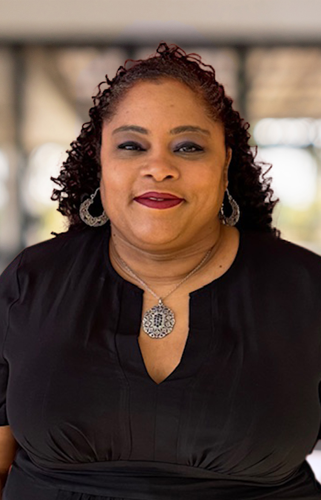 Danielle Joseph-Key, with curly dark hair, wearing a black dress, silver earrings, and a matching pendant necklace, smiles at the camera in a softly blurred indoor setting.