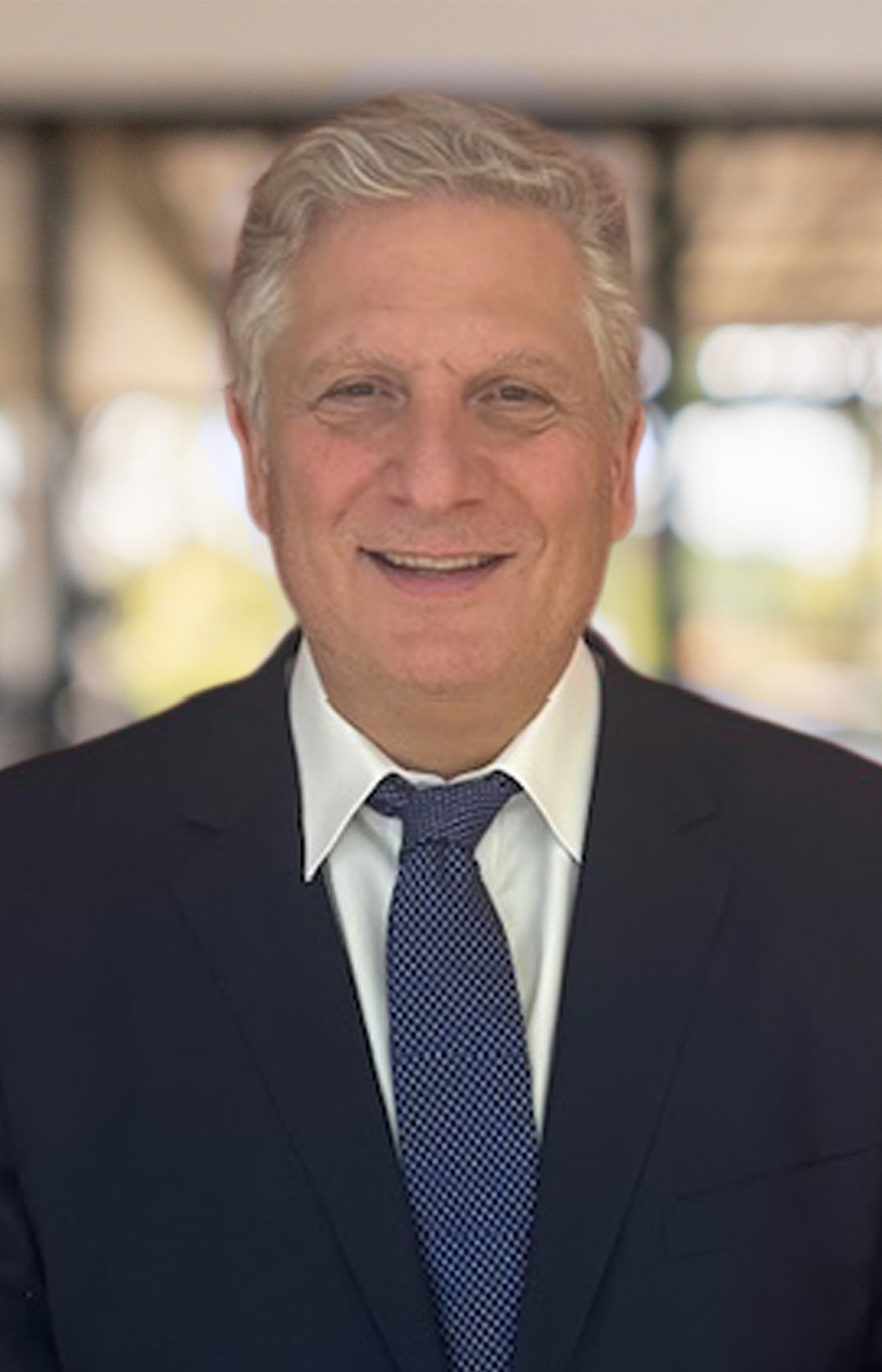 Dean T. Bennett, a smiling older man with gray hair, stands indoors in a dark suit, white shirt, and patterned tie against a blurred background.