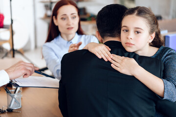 A young girl hugs a man tightly while a woman gestures and speaks to them across a desk with documents, suggesting a serious conversation about custody modified after divorce.