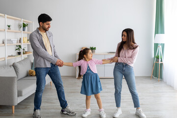 A young girl stands between a man and a woman, holding each of their hands as if the kid must choose who to live with. The adults face each other with serious expressions in a bright living room.