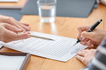 Close-up of two people’s hands as one points to a section on a document with a pen and the other signs it, with a glass of water and folders on a wooden table in the background.
