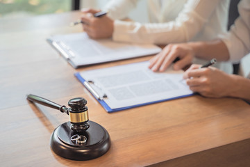 A wooden judge’s gavel and a pair of wedding rings rest on a table, while two people sit in the background, signing documents on clipboards, finalizing their divorce agreement modified to reflect new terms.