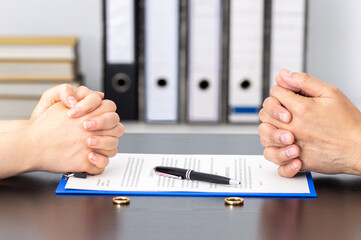 Two people sit across from each other with hands clasped, a pen and document between them on the table, and two wedding rings placed on the surface, suggesting a legal or divorce proceeding.