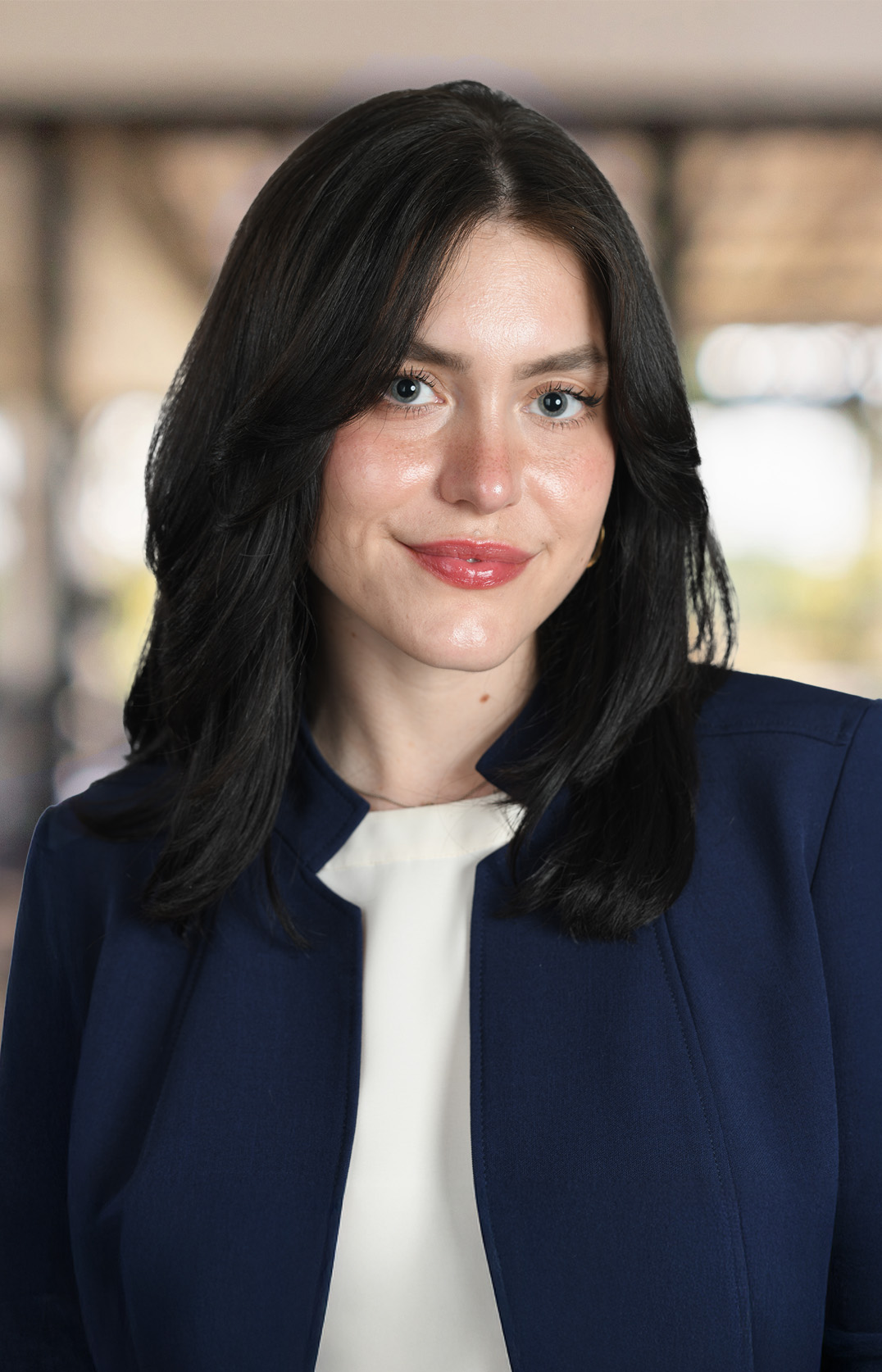 Wren G. Downs, a woman with long dark hair wearing a navy blazer and white top, smiles confidently at the camera, standing indoors with a blurred background.