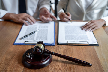 Two people sit at a table signing documents on clipboards, with a gavel and two wedding rings on a wooden surface in the foreground, symbolizing a New Jersey divorce, even if the couple was married in a different state.