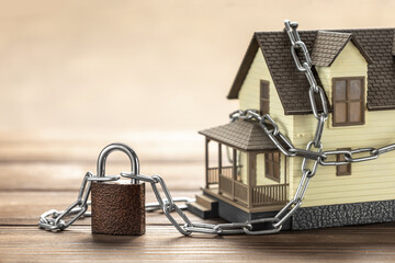 A model house wrapped in a metal chain secured with a padlock, symbolizing home security or property protection, set on a wooden surface with a neutral background—representing the impact of a lien or child support lien on property ownership.