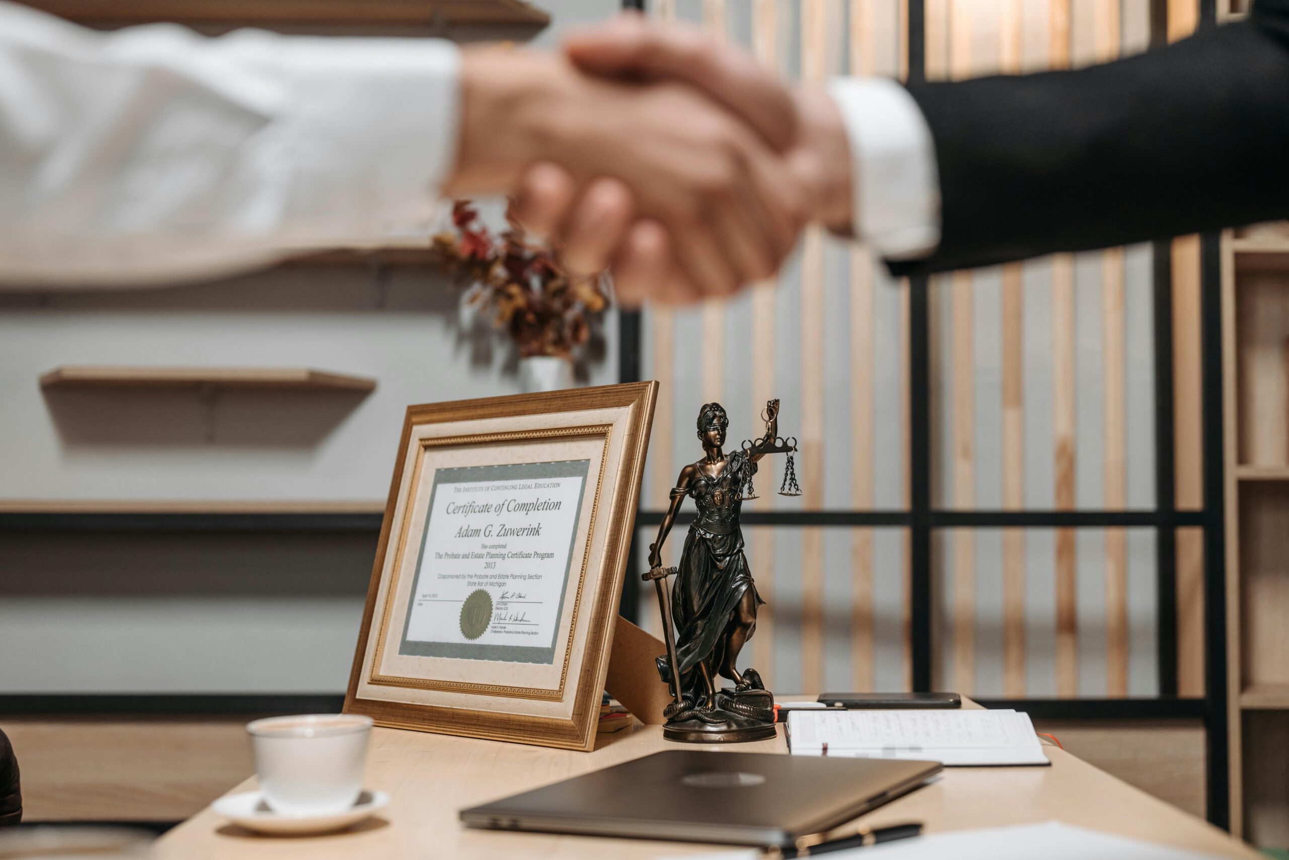 Two people in business attire shake hands over a desk with a framed certificate, Lady Justice statue, laptop, coffee cup, and documents—symbolizing how to file for divorce in New Jersey in a professional office setting.
