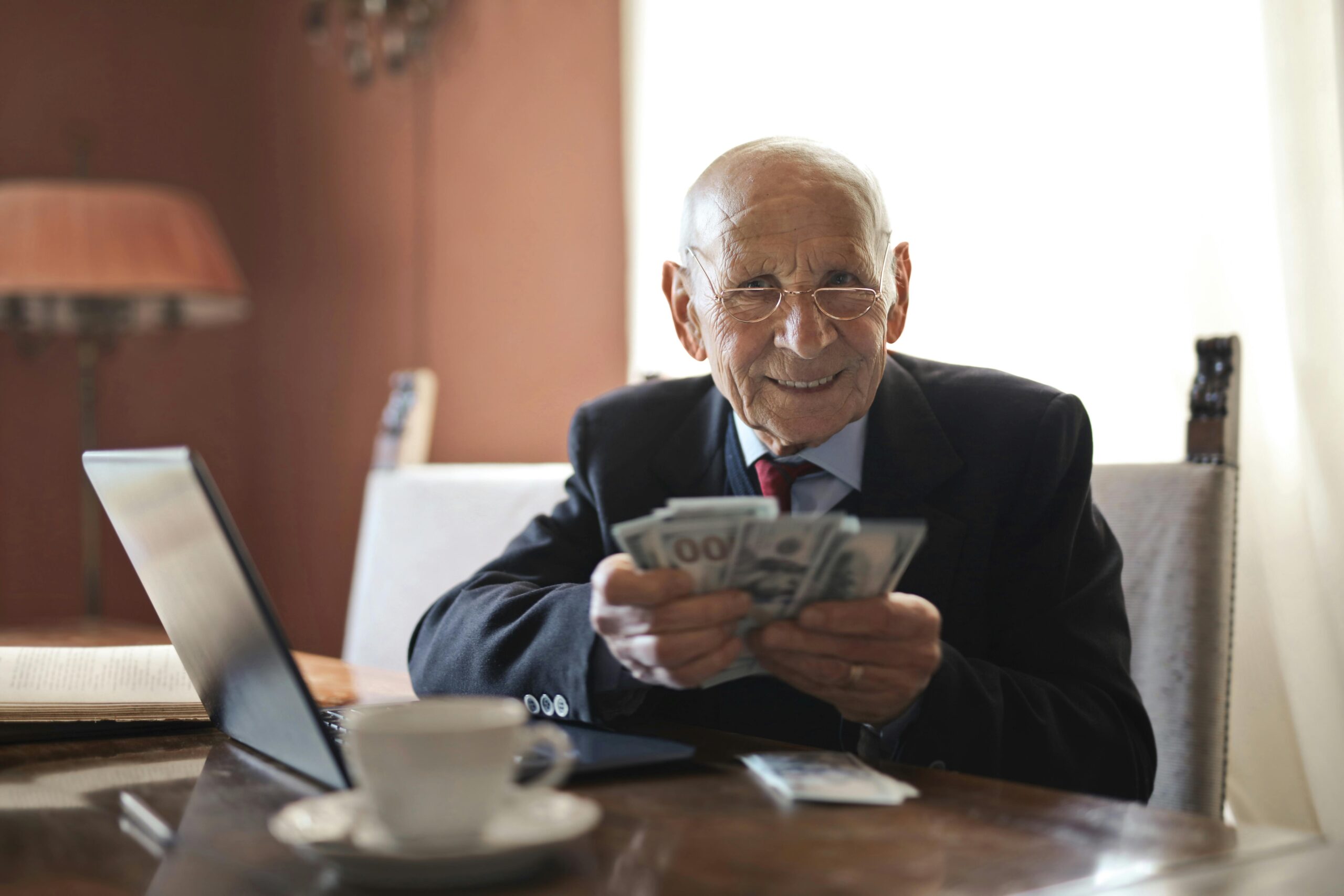 An elderly man in a suit sits at a table with a laptop, smiling and holding several 100-dollar bills—perhaps celebrating equitable distribution of retirement savings in New Jersey. A cup and saucer sit nearby, with a warm-toned room in the background.