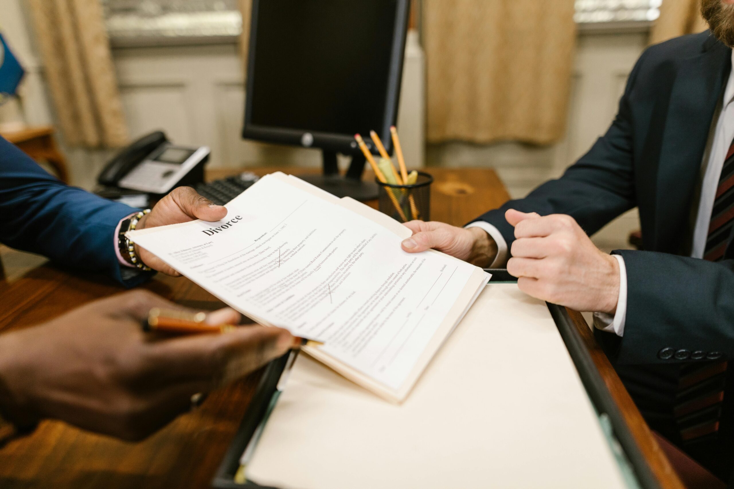 Two people in formal attire sit at a desk in New Jersey, exchanging and reviewing a finalized divorce document, with a computer monitor, pens, and papers visible in the background.