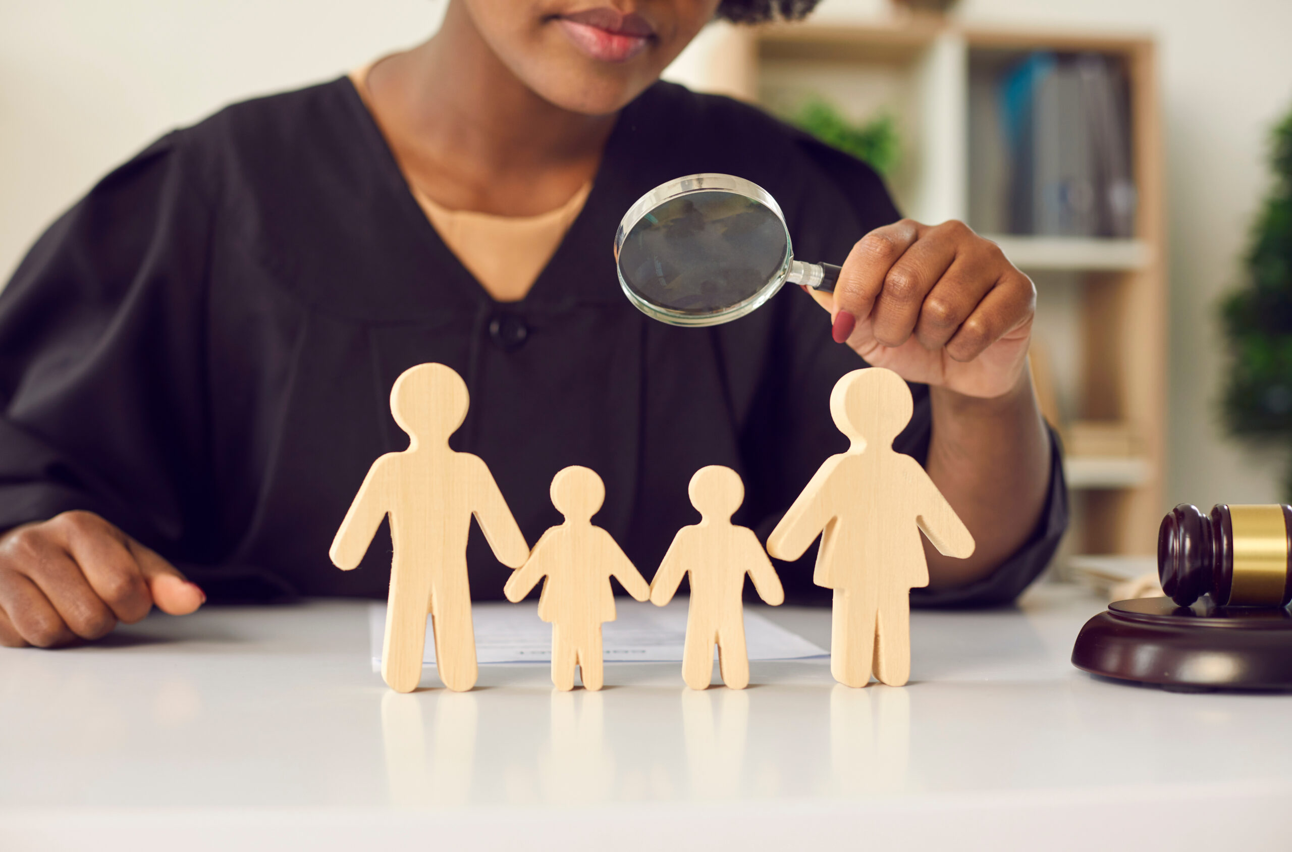 A judge in black robes closely examines wooden cutouts of a family with a magnifying glass. A gavel rests on the desk, symbolizing custody case proceedings in New Jersey, where a child may sometimes testify.