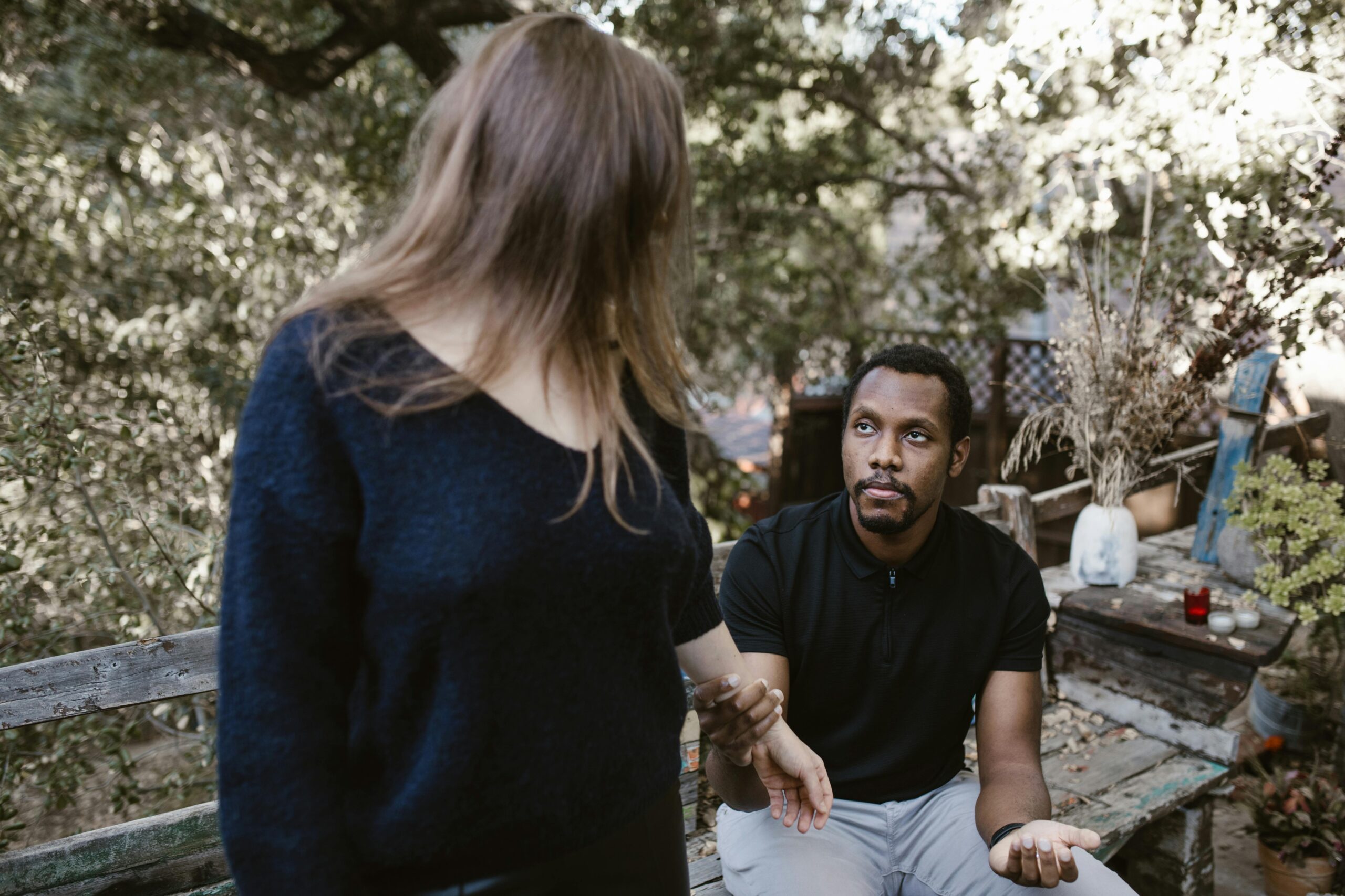 A man sits on a wooden bench outdoors, looking up seriously at a woman standing in front of him. Her back is to the camera, and the setting is surrounded by trees and plants, hinting at a tense discussion about New Jersey divorce and abandonment.