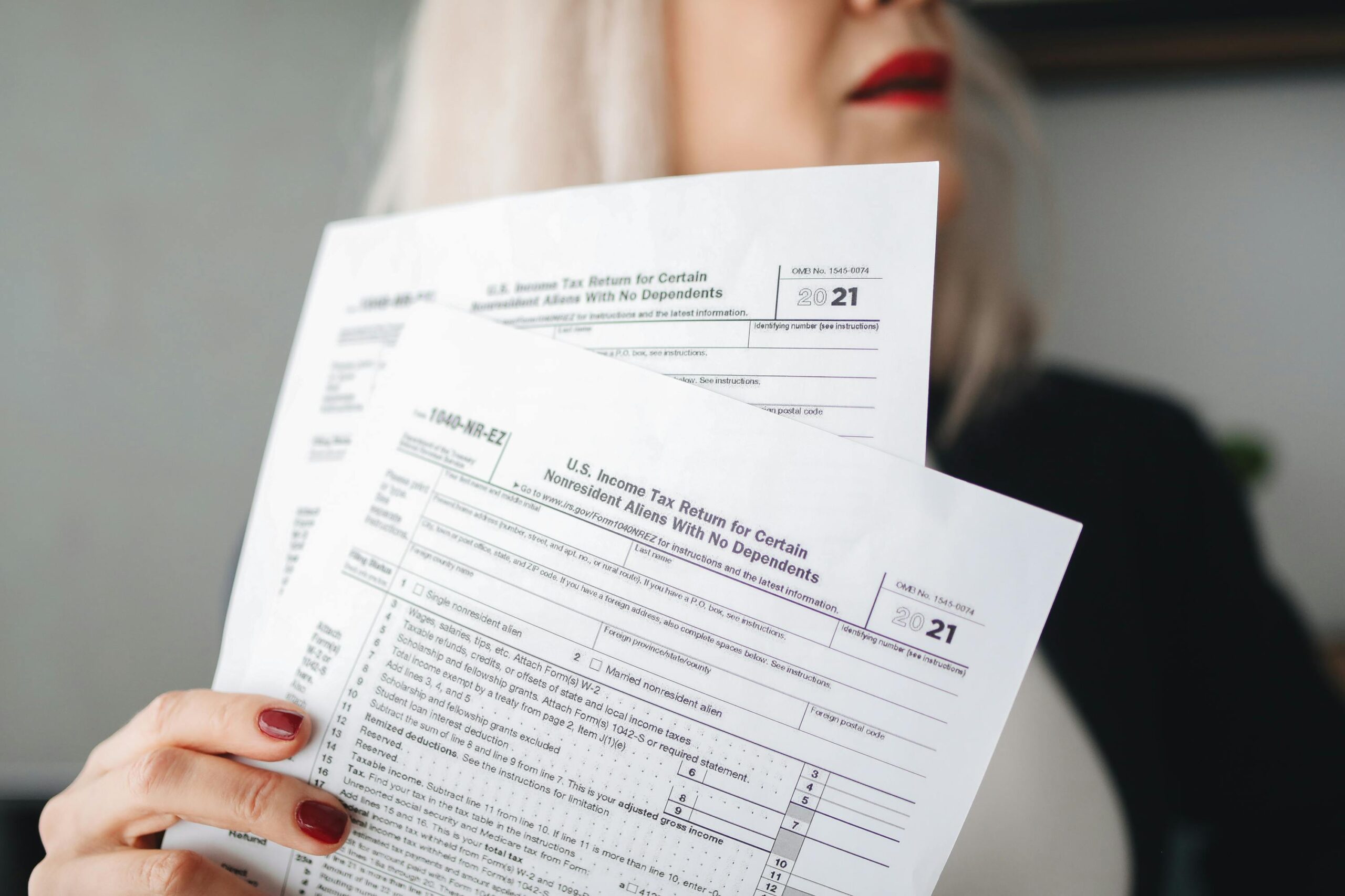 A woman with light hair holds up U.S. income tax return forms, partially visible, with the year 2021 printed on them. The focus is on the documents, with her face mostly out of frame.