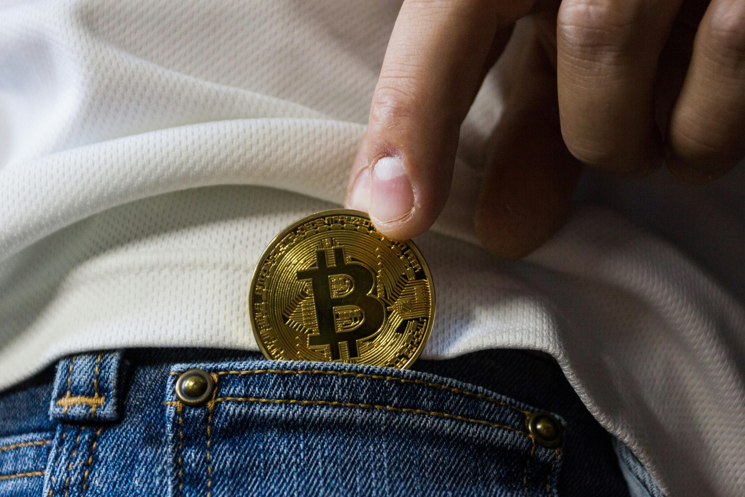 A person places a gold-colored Bitcoin coin into the front pocket of blue jeans, wearing a white shirt.